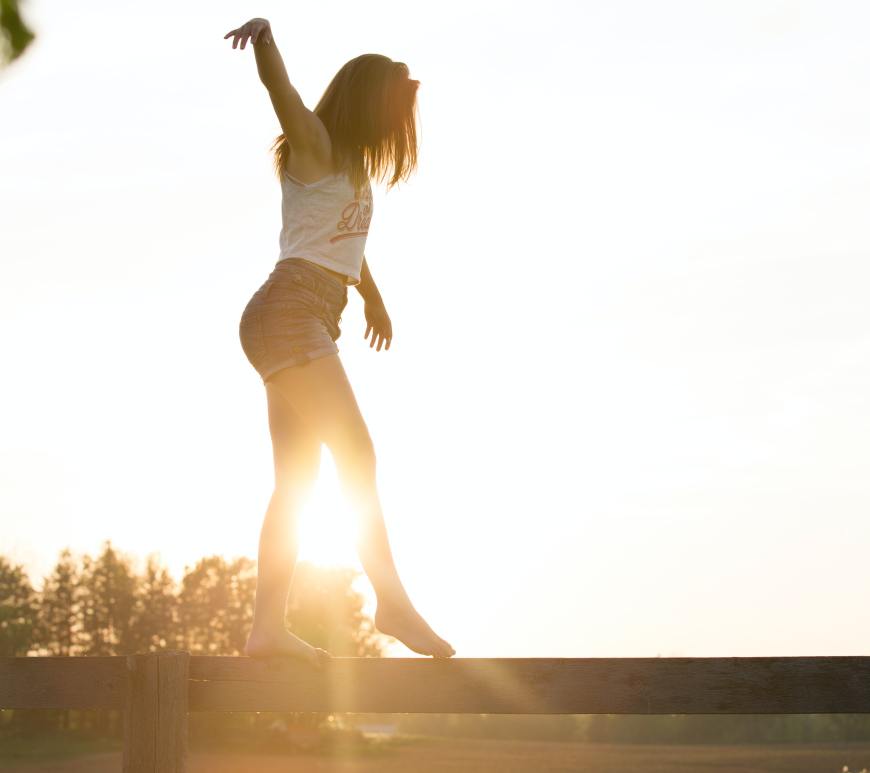 woman in a tank top and shorts walking across a wooden fence like a balance beam with the sun and trees in the background