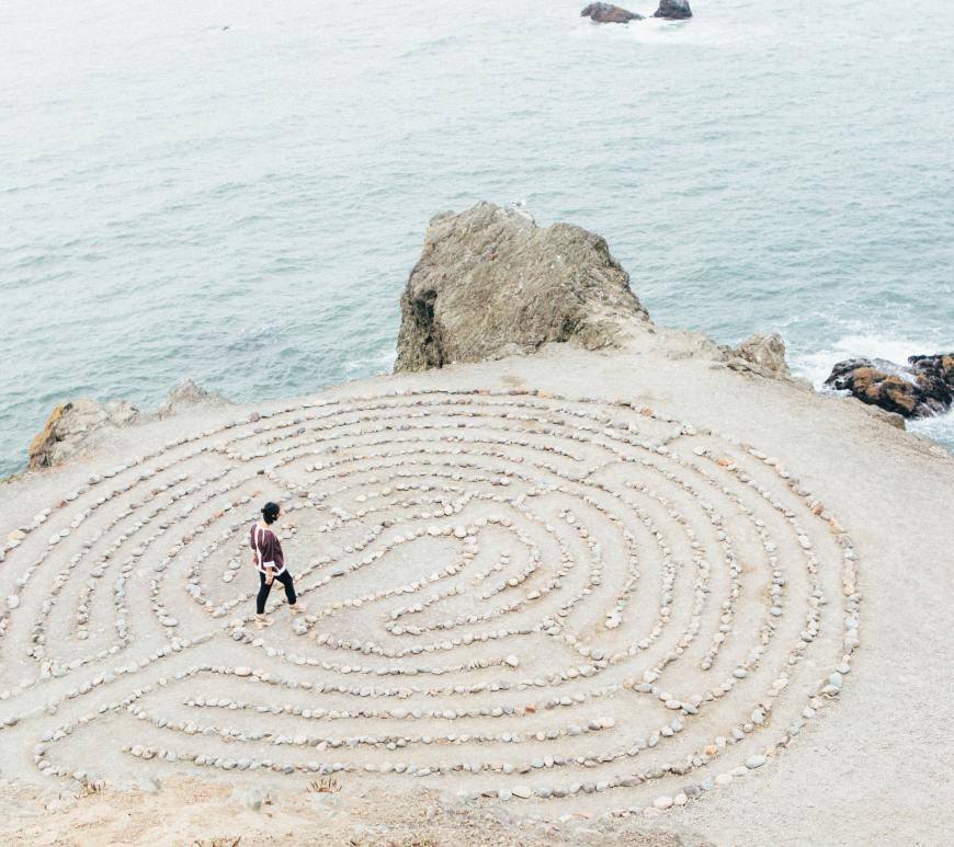 person walking through a circular stone maze in the sand by the ocean