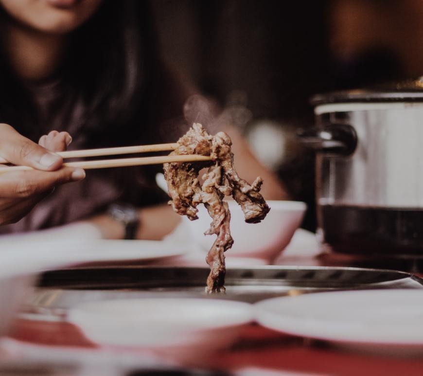 woman holding meat using chopsticks over table with plates and crockpot